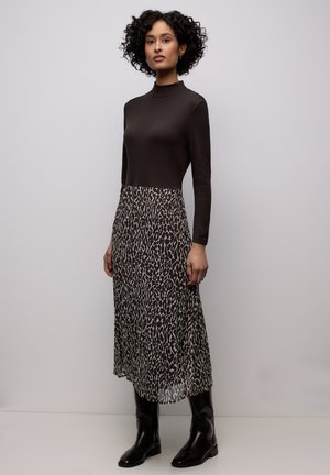 Frau mit lockigem Haar, die ein schwarzes Oberteil mit hohem Kragen, einen Midi-Rock mit Animal-Print und schwarze kniehohe Stiefel trägt, steht vor einem einfarbigen Hintergrund.