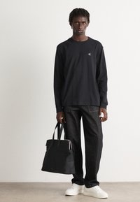 Ung mann i svart langermet skjorte, mørke jeans og hvite sneakers står med en svart totebag mot en enkel hvit vegg.