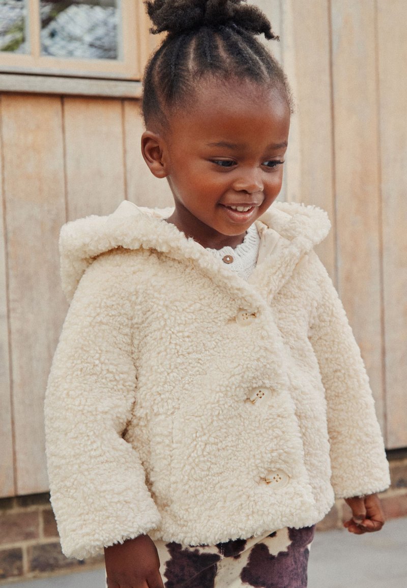 Veste en shearling duveteuse de couleur crème avec capuche et trois boutons, associée à un pantalon imprimé à motifs marron et blanc.