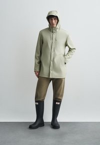 Jasnozielona wodoodporna kurtka z kapturem, zatrzaskami i bocznymi kieszeniami, zestawiona z brązowymi krótkimi spodniami i czarnymi gumowymi butami.