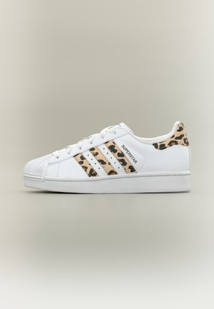 Bílá teniska Adidas Superstar s leopardím potiskem na třech pruzích a patce, plochá podrážka a tkaničky na neutrálním pozadí.