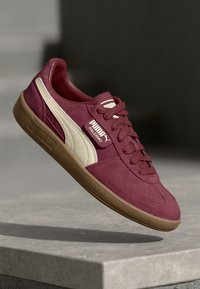 Bordowe zamszowe sneakersy z kremowymi akcentami, teksturowaną gumową podeszwą i wyraźną metką z logo na języku. Klasyczny zaokrąglony kształt.