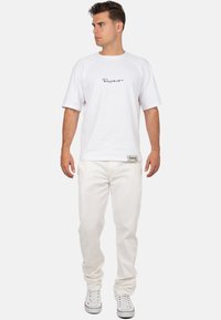 Witte katoenen T-shirt met korte mouwen, voorzien van een zwart handgeschreven logo. Gecombineerd met witte broek en klassieke lage sneakers.