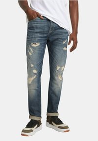 Jeans in denim blu sbiadito con segni di usura e toppe sfrangiate, abbinati a sneaker multicolori con suola bianca e accenti neri.