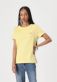 Tricou din bumbac galben cu decolteu rotund și mâneci scurte. Un mic logo "BOSS" este brodat în auriu. Purtați cu blugi albaștri.
