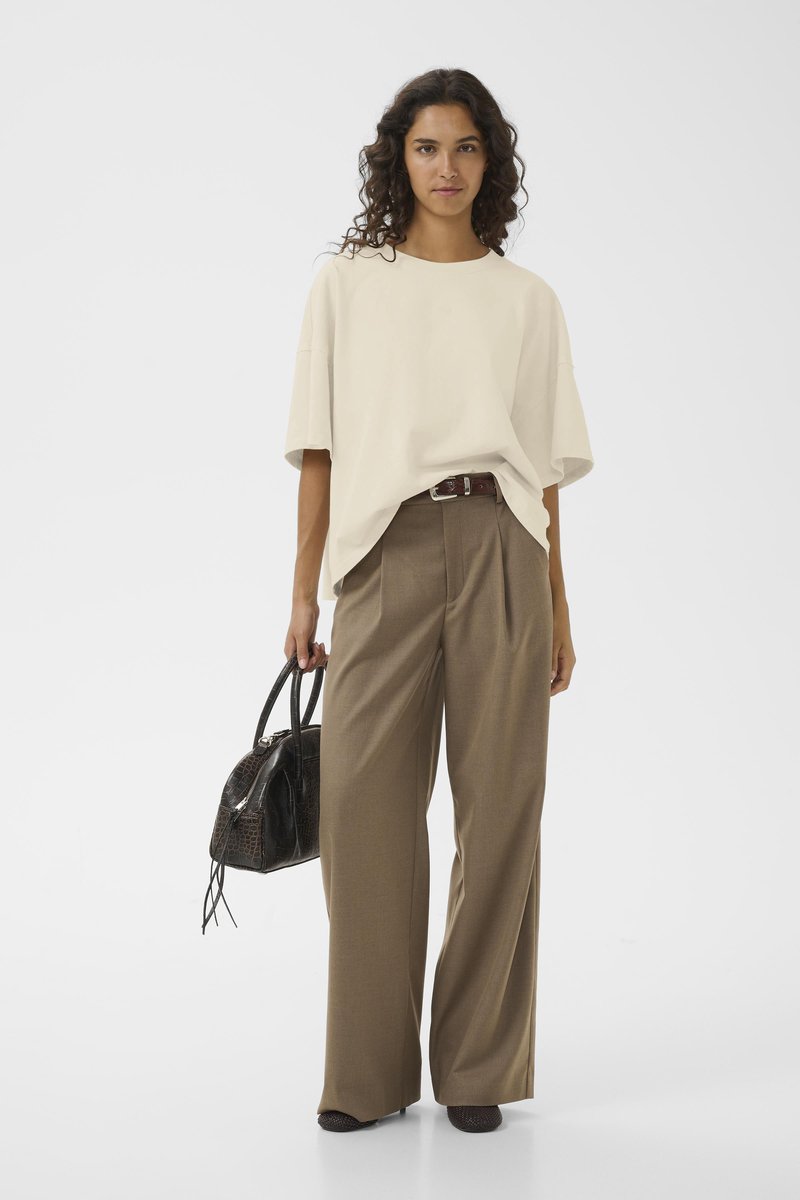 Beige übergroßes T-Shirt kombiniert mit hoch taillierten, weit geschnittenen braunen Hosen. Das Modell hält eine schwarze, strukturiert Handtasche.
