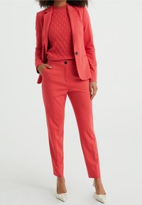 Coral blazer and matching slim-fit trousers over a textured coral sweater. White heeled shoes. Features classic lapels and front pockets.