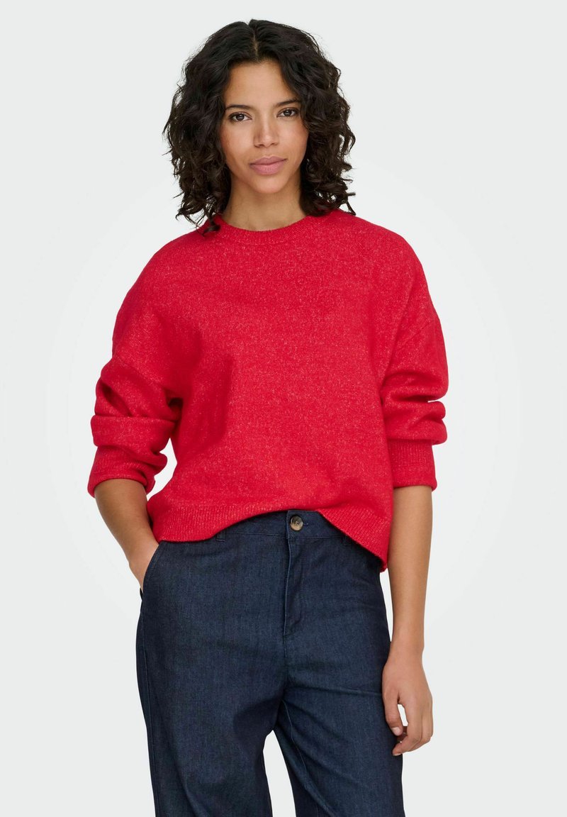 Pull en maille rouge avec un col rond et manches retroussées, associé à un jean bleu foncé. Le pull présente une texture douce et une coupe décontractée.