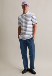 Gestreiftes T-Shirt in Weiß und Navy, mit Rundhalsausschnitt und kurzen Ärmeln. Kombiniert mit blauen Hosen und schwarzen Schuhen. Lavendelfarbenes Cap mit Logo.