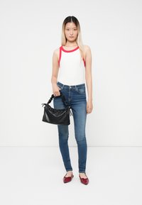 Bodysuit blanco con ribete rojo, jeans ajustados azules, bolso negro texturizado y zapatos planos rojos. La modelo se encuentra de pie frente a un fondo blanco.