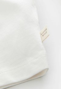 T-shirt bianco in cotone con un piccolo etichetta di tessuto con la scritta "Little Bird" stampata, visibile sull'orlo. Texture liscia e design semplice.