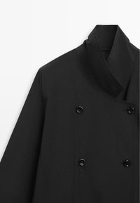 Zwarte dubbelgeknoopte jas met hoge kraag, zichtbare stiksels en vier zwarte knopen aan de voorkant, tentoongesteld tegen een witte achtergrond.