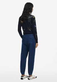 Suéter negro con bordado floral colorido, combinado con joggers de mezclilla azul con puños elásticos y dos bolsillos traseros. Sandalias blancas.