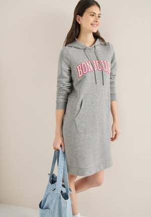 Femme portant une robe à capuche grise avec "BONJOUR" en lettres roses, tenant un sac fourre-tout bleu clair, debout devant un fond beige.