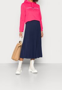 Roze hoodie met logo, navy plissérok, witte enkellaarsjes en een beige totebag. Focus op stoftexturen en kleurcontrasten.