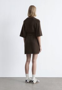 Braun gestricktes Kleid mit kurzen Ärmeln, tailliert und mit geripptem Saum. Kombiniert mit weißen Socken und Schuhen mit marineblauen Streifen.