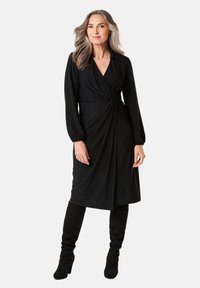 Schwarzes Wickelkleid mit langen Ärmeln, drapierter Gestaltung und V-Ausschnitt. Getragen mit kniehohen schwarzen Stiefeln. Glattes Material und gerafftes Detail an der Taille.