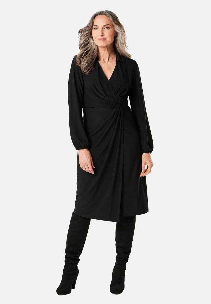 Schwarzes Wickelkleid mit langen Ärmeln, drapierter Gestaltung und V-Ausschnitt. Getragen mit kniehohen schwarzen Stiefeln. Glattes Material und gerafftes Detail an der Taille.