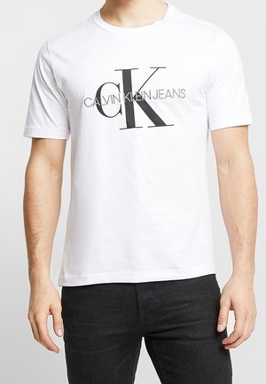 T-shirt en coton blanc avec un grand logo noir "CK" et le texte "CALVIN KLEIN JEANS" dans une police moderne. Design à manches courtes et encolure ras du cou.