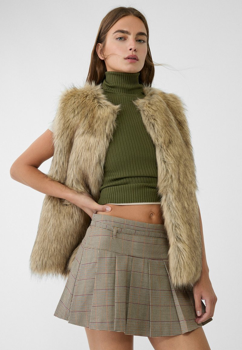 Gilet en fausse fourrure marron clair avec un crop top vert à côtes et une jupe plissée à carreaux comportant des lignes rouges et noires.