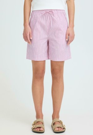 Person iført lyserøde og hvide shorts med lodrette striber, snøre i taljen og beige sandaler, stående foran en ensfarvet baggrund.