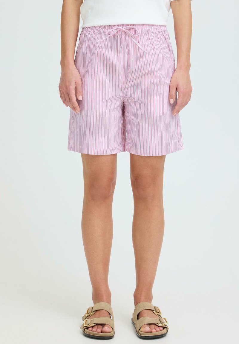 Άτομο που φοράει ριγέ σορτς σε ροζ και λευκό με κορδόνι στη μέση και μπεζ σανδάλια, στέκεται μπροστά σε απλό φόντο.