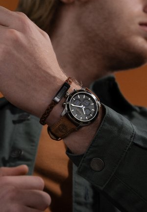 Orologio cronografo nero con accenti argento e cinturino in pelle marrone, abbinato a un bracciale intrecciato marrone. Sfondo arancione.