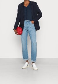 Person klädd i marinblå kavaj, mörkblå knäppt topp, ljusblå uppvikta jeans, vita sneakers, som håller en röd kuvertväska mot en enfärgad bakgrund.