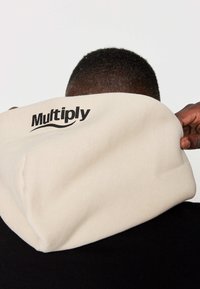 Sudadera con capucha beige con un gran logotipo negro "Multiply" en la parte posterior, hecha de tela suave, llevada con una capa base negra.