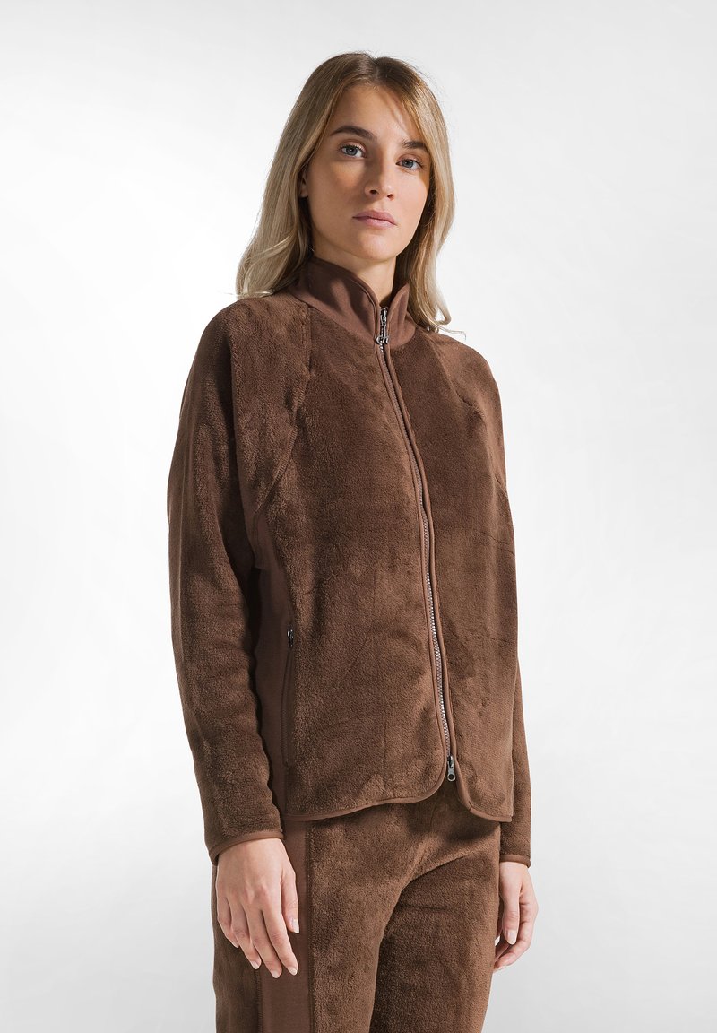 Giacca in pile marrone con colletto alto e chiusura con zip. Presenta una consistenza morbida e tasche sui lati. Abbinata a un pantalone in pile coordinato.