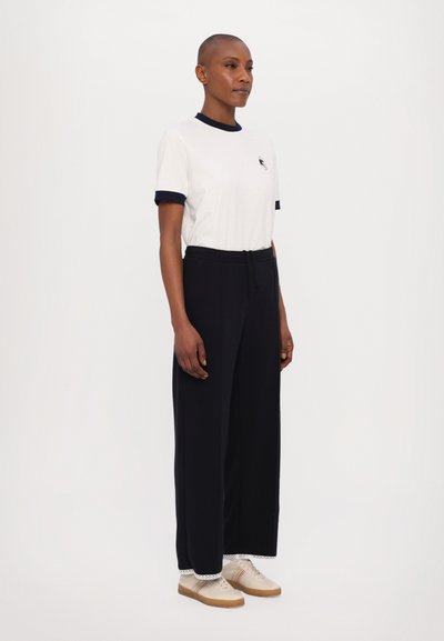 White short-sleeve t-shirt with navy blue trim, paired with black wide-leg pants and light shoes featuring beige and striped accents.