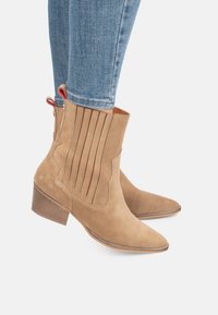 Botas de tobillo de ante en color beige con puntera afilada, presentan pliegues verticales en el lateral, un tacón bajo en bloque y una tira decorativa para facilitar la colocación.