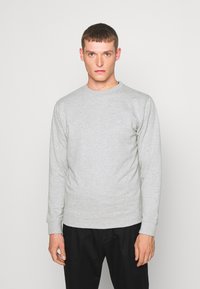 Grå bomulds sweatshirt med lange ærmer, rund hals og ribbede mansjetter. Har et diskret logo på brystet og bæres med sorte bukser.