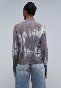 Maglione tie-dye marrone e grigio con colletto alto, maniche lunghe e vestibilità rilassata, abbinato a jeans azzurri chiari. Morbido al tatto.