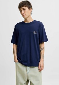 Granatowa bawełniana koszulka z luźnym krojem, krótkimi rękawami i małym białym detalem logo po lewej stronie. Noszona z jasnymi spodniami.