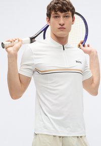 Άσπρο κοντομάνικο πόλο πουκάμισο με φερμουάρ στο γιακά, διακοσμημένο με μαύρες και χρυσές οριζόντιες ρίγες. Φορώντας το, ένα μοντέλο κρατά ρακέτα τένις.