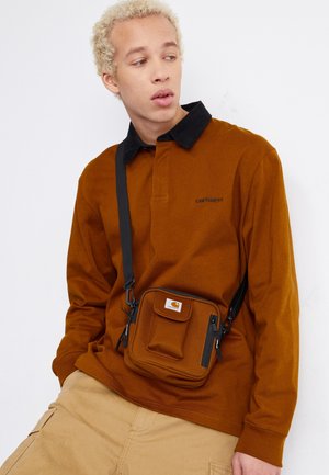 Ung person med kort blondt hår iført en brun langærmet skjorte og crossbody taske, der kigger til siden mod en hvid baggrund.