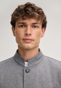 Grijze herringbone jas met een opstaande kraag, éénknopsluiting en textiel met structuur. Witte blouse eronder, zonder zichtbare patronen.
