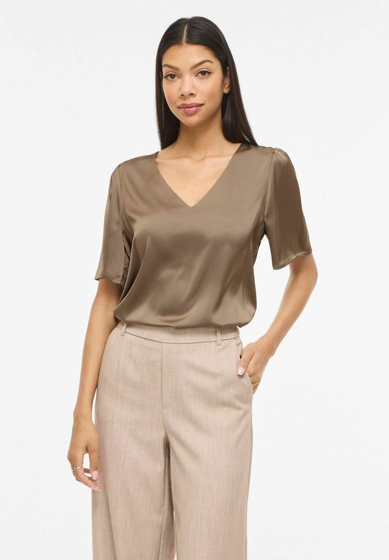 Femme portant un chemisier en satin marron à manches courtes et un pantalon beige taille haute, posant avec une main dans la poche sur fond blanc.