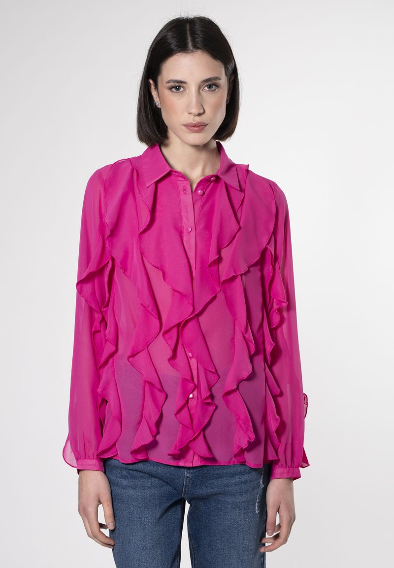 Blusa in chiffon rosa con design a volant, chiusura a bottoni e maniche lunghe. Presenta una consistenza trasparente e dettagli a strati. Indossata con jeans blu.