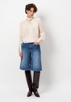 Beige Seidenbluse mit hohem Kragen, blaue Denim-Culottes und braune, krokodilprimitierte kniehohe Stiefel. Das Model steht mit einer Hand in der Tasche.