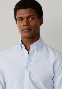 Uomo con capelli corti e ricci che indossa una camicia azzurra con bottoni, che guarda leggermente verso destra contro uno sfondo semplice.