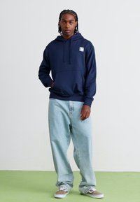 Navy blue hooded sweatshirt with a front pocket and logo, paired with light blue baggy jeans and gray sneakers on a green floor.
