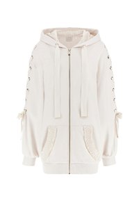 Light beige zip-up hoodie with a lined hood, lace-up accents on sleeves, ruffled pocket edges, and tie details on cuffs. Soft, textured fabric.