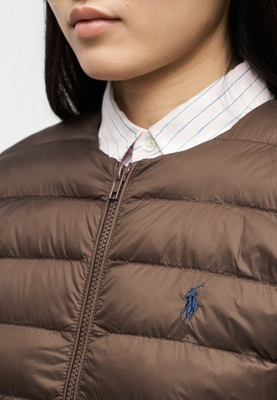 Γυναίκα που φοράει καφέ μπουφάν με ραφές και φερμουάρ μπροστά, και πουκάμισο με μπλε ρίγες από κάτω.