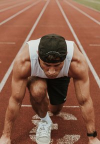 Homme athlétique en débardeur blanc et shorts noirs sur une piste rouge, portant une casquette noire avec "CONTINUE" et des baskets blanches.