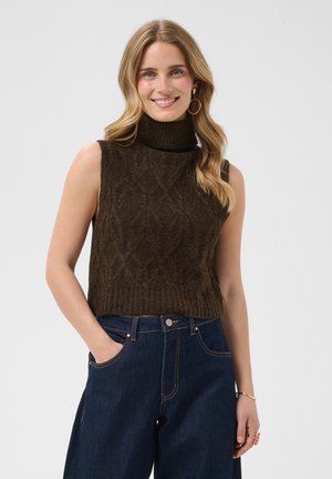 Smilende kvinde med bølget blondt hår iført store hoop-øreringe, ærmeløs mørkebrun strikket højhalset top og mørkeblå højttaljede jeans.