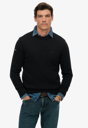 Pull en tricot noir avec des poignets côtelés, superposé à une chemise en denim bleu clair, associé à un jean bleu et une ceinture marron.