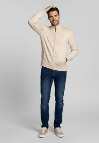 Suéter crema con cremallera y cuello alzado, usado sobre jeans azules y zapatillas beige. Presenta puños acanalados y un logotipo minimalista.