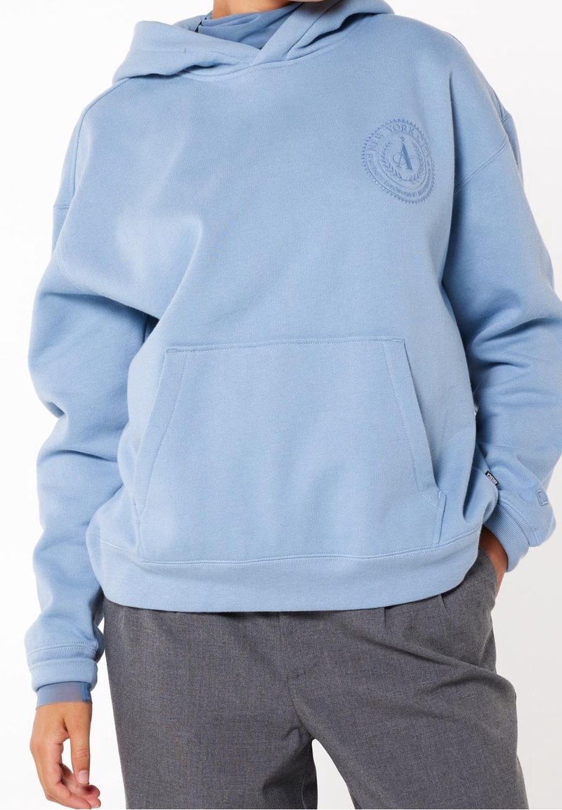 Personne portant un sweat à capuche bleu clair avec une poche avant et un logo circulaire en relief sur la poitrine, associée à un pantalon gris, les mains détendues le long du corps.